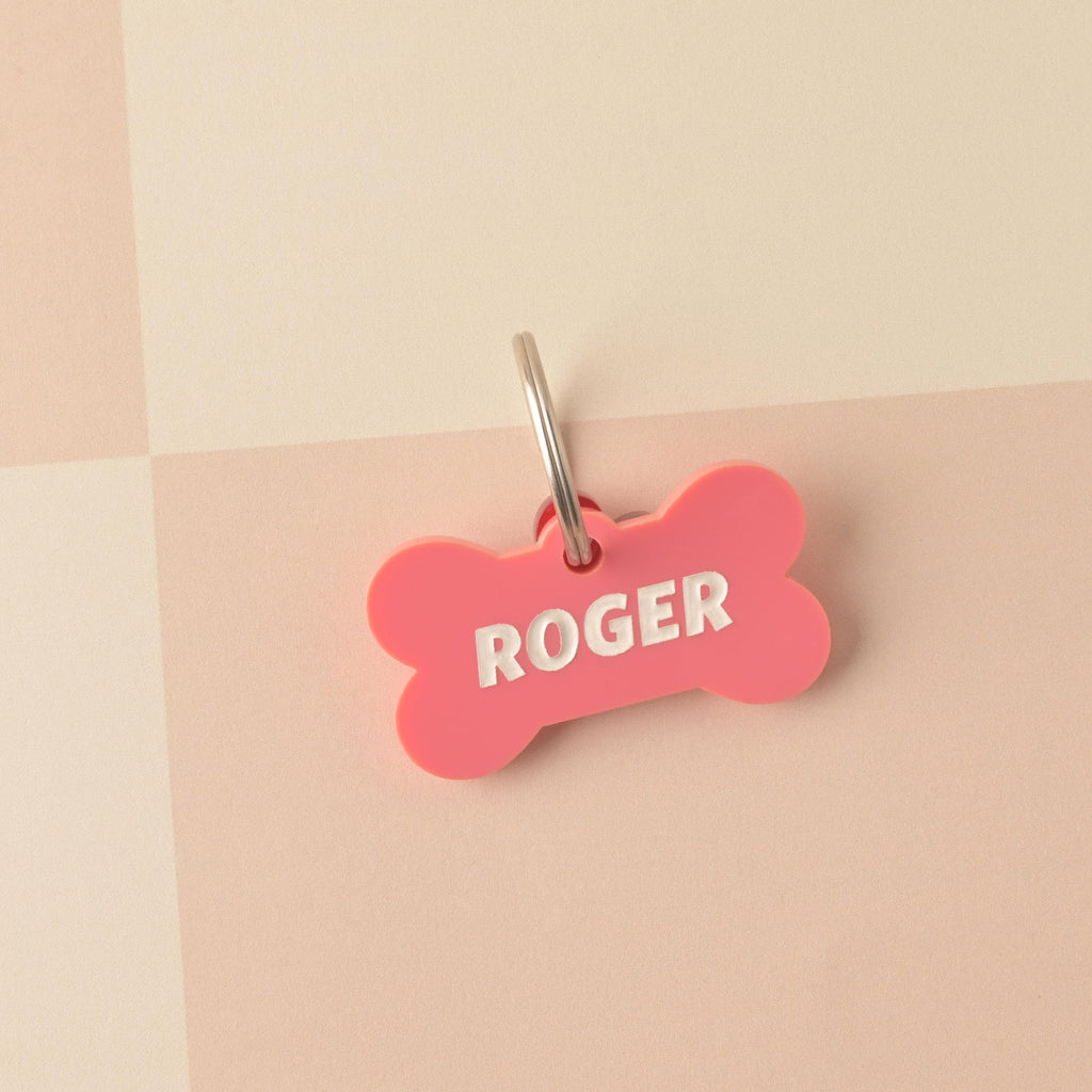 Close-up of a pink bone-shaped acrylic pet ID tag, personalized with the name 'ROGER' engraved in large white text. A silver split ring is attached. The tag is displayed on a light pink and white checkered background. Bones Day, Bone shaped Dog Tag, Personalized Pet Tag, Cat and Dog ID Tag