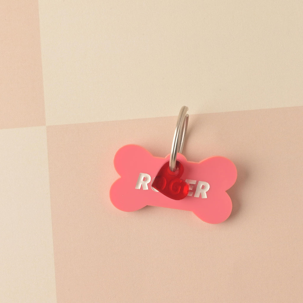 Close-up of a pink bone-shaped acrylic pet ID tag personalized with the name 'ROGER' in silver text. A small, translucent red heart charm is attached to the silver split ring. The tag is displayed on a light pink and white checkered background. Bones Day, Bone shaped Dog Tag, Personalized Pet Tag, Cat and Dog ID Tag