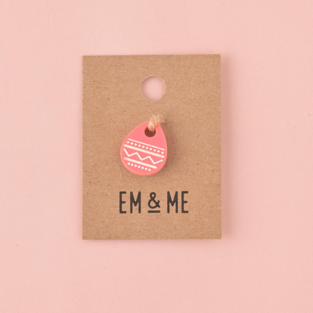 A single bright pink acrylic charm shaped like an Easter egg, decorated with white engraved patterns. The charm is displayed on a small square kraft paper backing card with the EM & ME logo printed below it, set against a solid pink background.