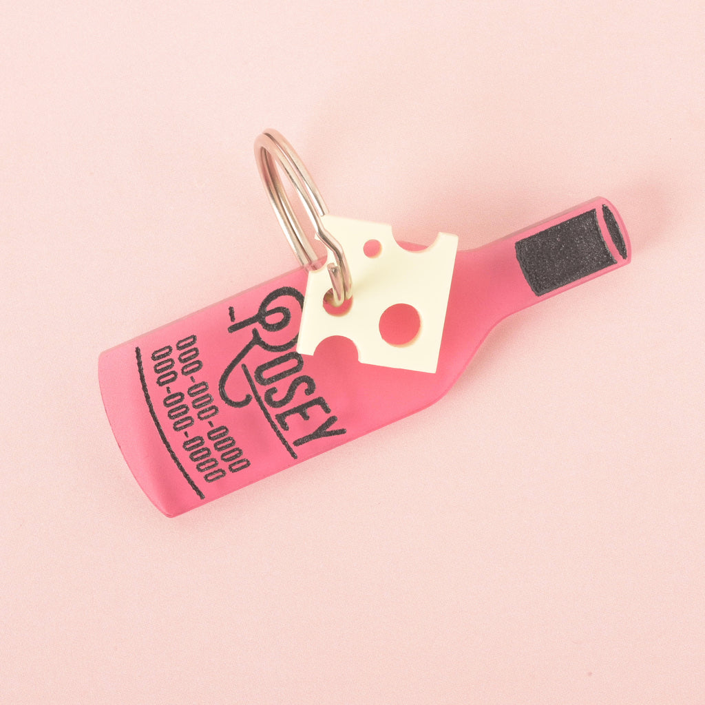 A pink wine bottle-shaped pet tag with the name "ROSEY" and a placeholder phone number on it. The tag is attached to a small, white cheese charm. The items are connected to a silver ring, perfect for a collar, and are shown on a light pink background.