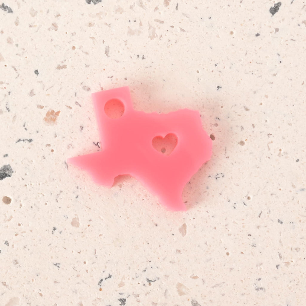 Pink acrylic Texas state-shaped charm with a heart cutout and top hole for hanging, displayed on a speckled cream terrazzo background.
