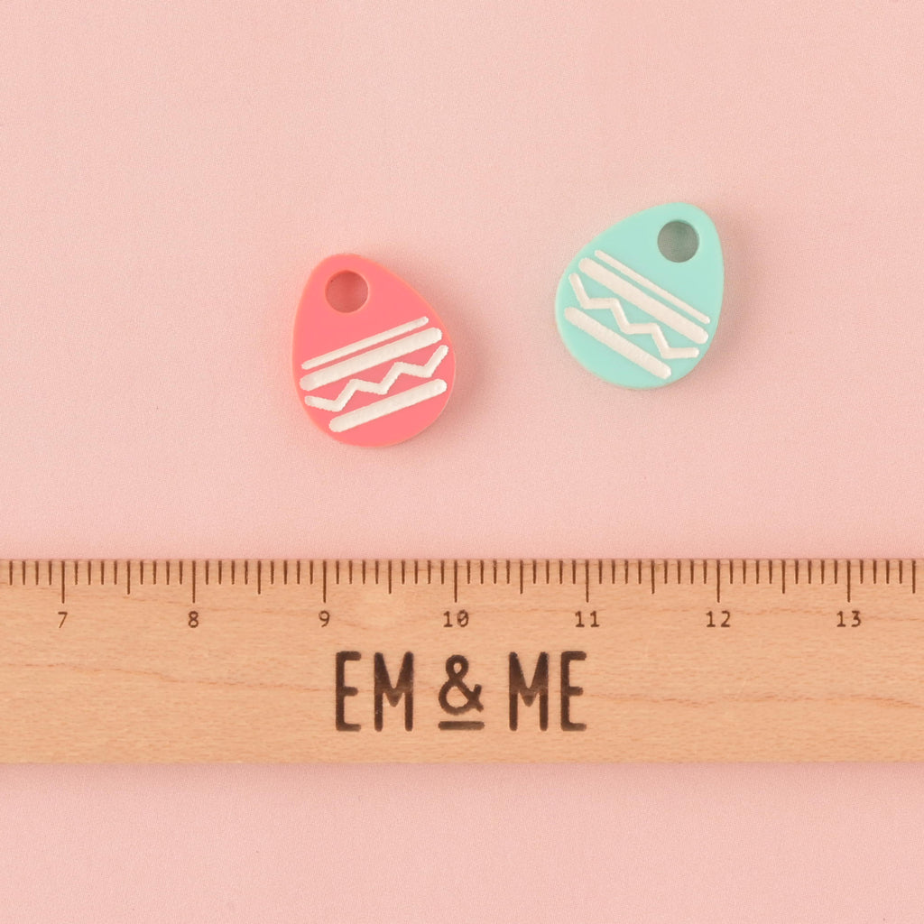 Two small acrylic Easter egg charms, one pink and one mint green, both decorated with white zigzag and stripe patterns. The charms are displayed above a wooden ruler marked with the brand EM & ME, on a light pink background.