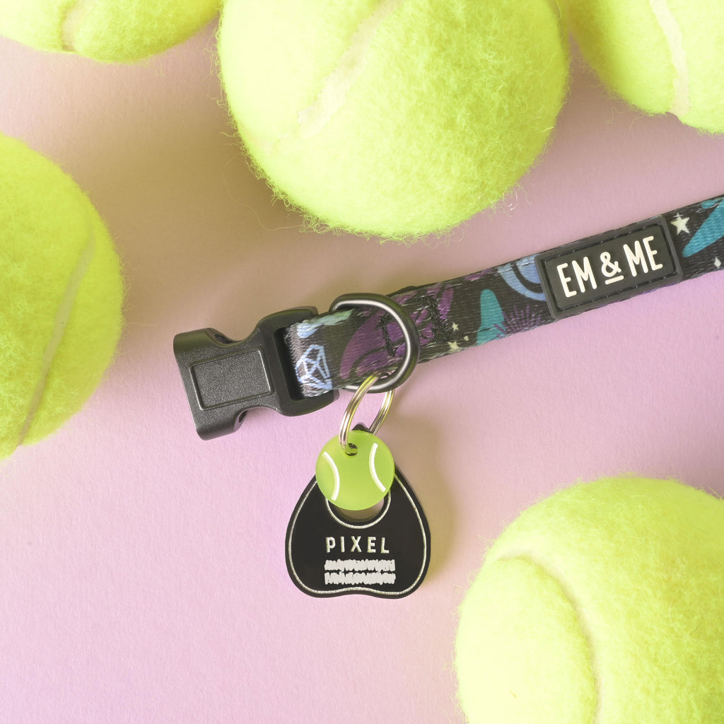 A personalized black Ouija planchette-shaped dog tag with the name "PIXEL" attached to a wide, patterned collar that reads "EM & ME." A small green tennis ball charm is also attached to the tag's ring. The items are surrounded by bright yellow-green tennis balls on a solid pink background.