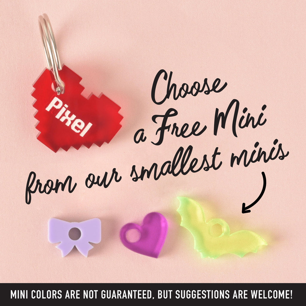 A xs. red pixelated heart pet tag that says "Pixel," a purple bow charm, a pink heart charm, and a translucent yellow bat charm are shown. The image also contains the text "Choose a Free Mini from our smallest minis" and "MINI COLORS ARE NOT GUARANTEED, BUT SUGGESTIONS ARE WELCOME!" The items are displayed on a solid pink background.