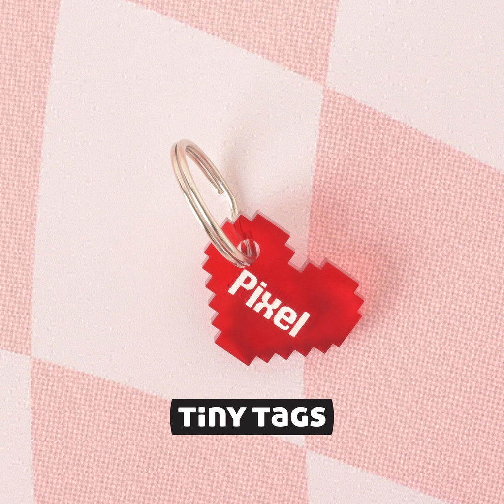 A close-up shot of a small, red pixelated heart pet tag that says "Pixel," attached to a silver ring. The tag is resting on a pink and white checkered surface. The image also contains the text "TINY TAGS" at the bottom.