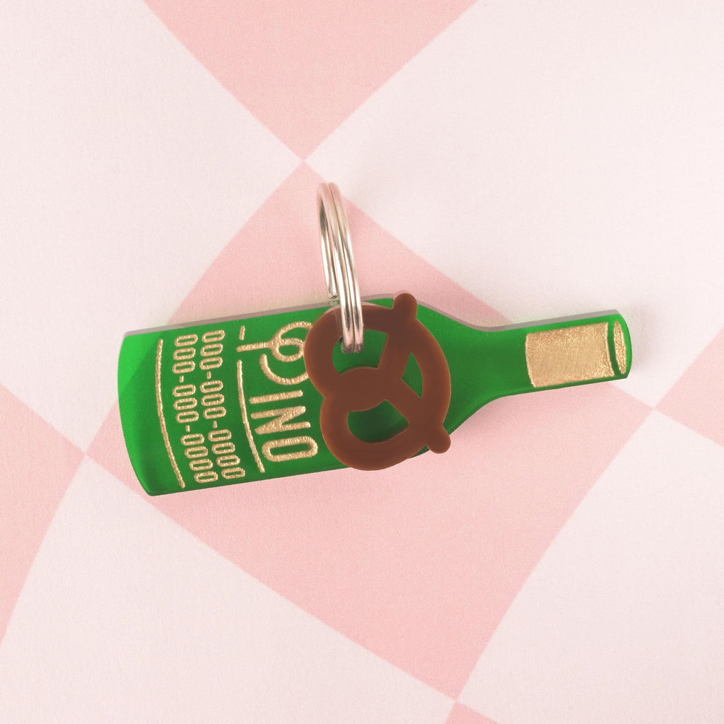 A personalized green wine bottle-shaped pet tag with the name "Pino" and a placeholder phone number. A small, brown pretzel charm is attached to the tag. Both are connected to a silver ring for a collar and are displayed on a pink and white checkered background.