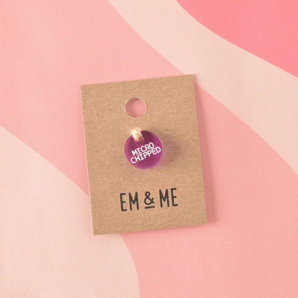 A small, circular, translucent purple acrylic charm engraved with "MICROCHIPPED" in white text. The charm is secured with twine to a branded 'EM & ME' kraft card.