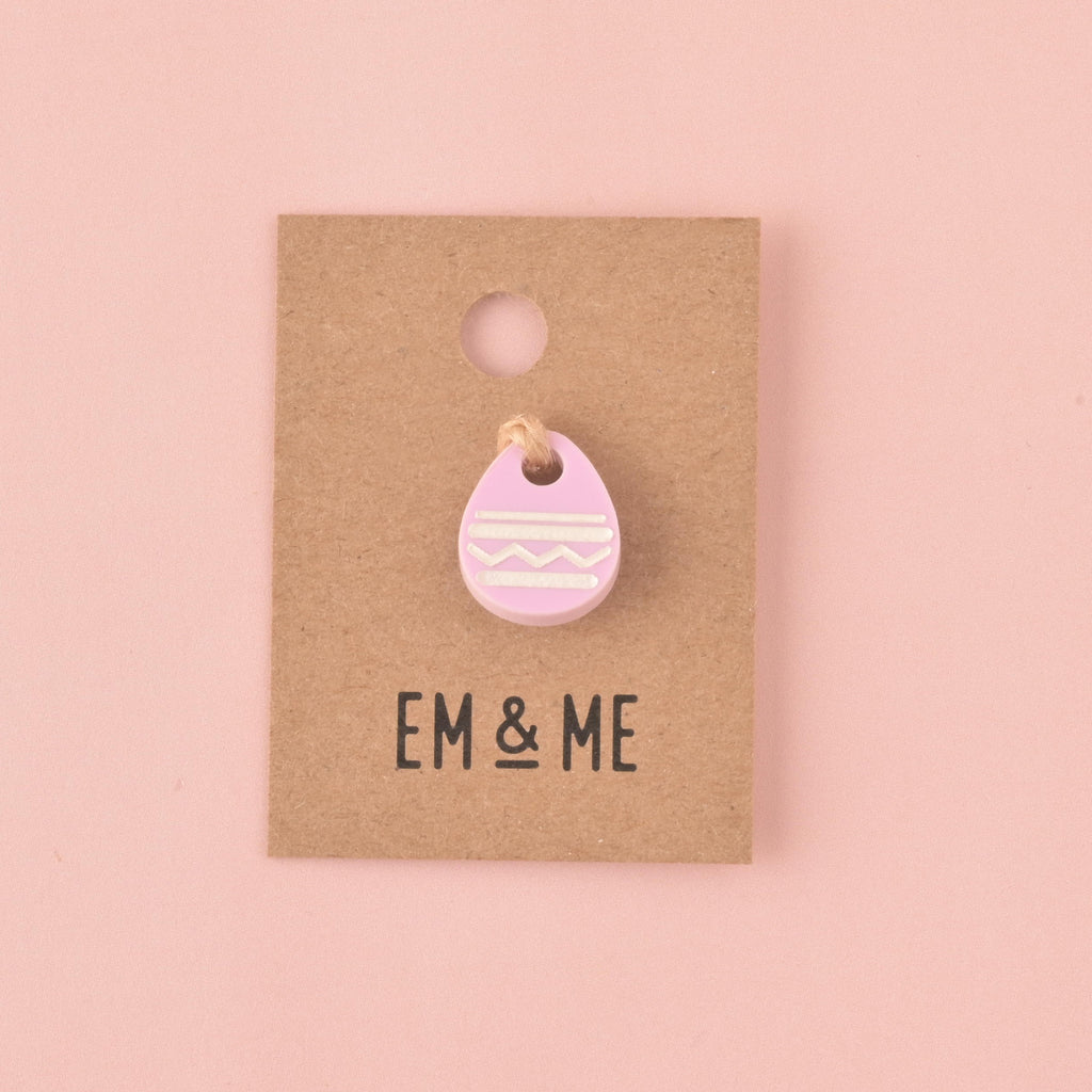 A single light purple (lilac) acrylic charm shaped like an Easter egg, decorated with white engraved patterns. The charm is displayed on a small square kraft paper backing card with the EM & ME logo printed below it, set against a solid pink background.