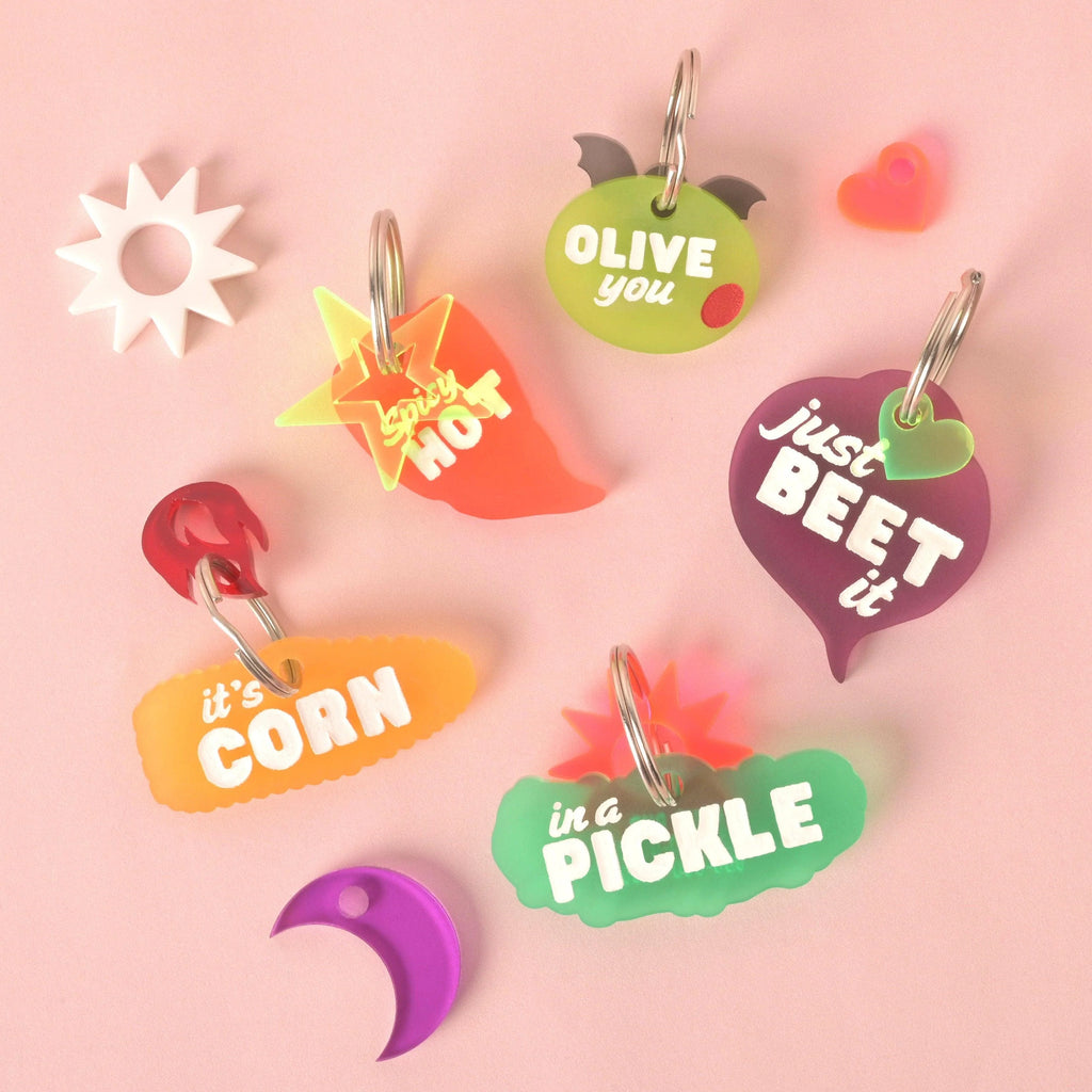 A collection of five humorous, food-themed acrylic pet ID tags: a green olive shape ('Olive You'), an orange chili pepper ('Spicy Hot') with a yellow star charm, a pickle shape ('In a Pickle') with a pink sunburst, an orange corn shape ('It's Corn'), and a purple beet shape ('Just Beet It'). Assorted charms (white star, heart, moon) are scattered around the tags. Spicy Hot, Pepper Personalized Pet Tag, ID for Cats and Dogs, Veggie Pet Tags