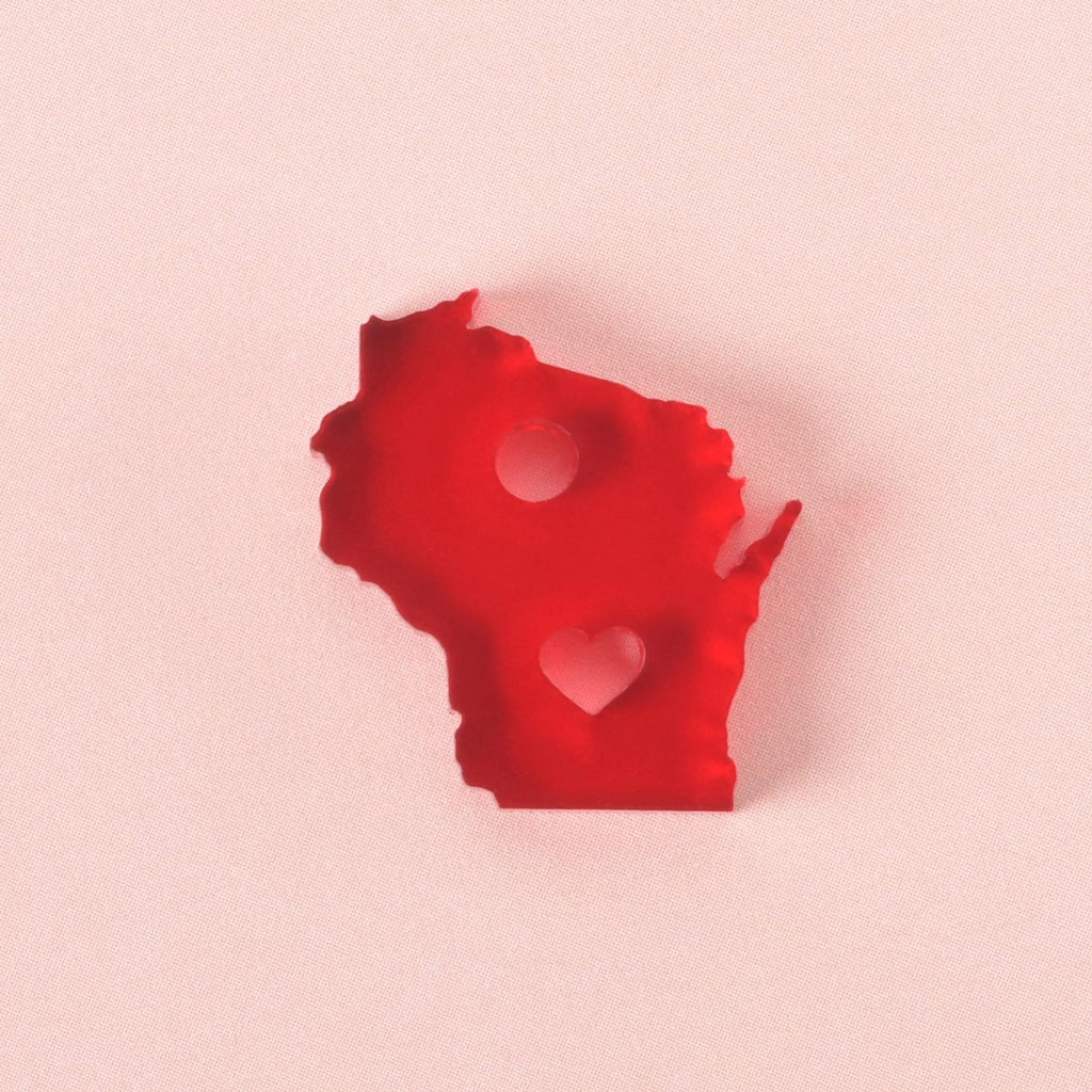 Close-up of red acrylic Wisconsin state charm with heart cutout and hanging hole, placed on a soft pink background.