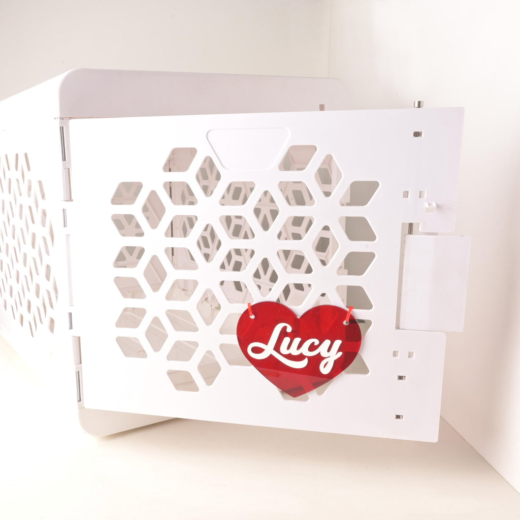 Custom red plastic heart-shaped pet ID crate tag with the name Lucy in white script, hanging on the door of a modern white pet crate with geometric cutouts