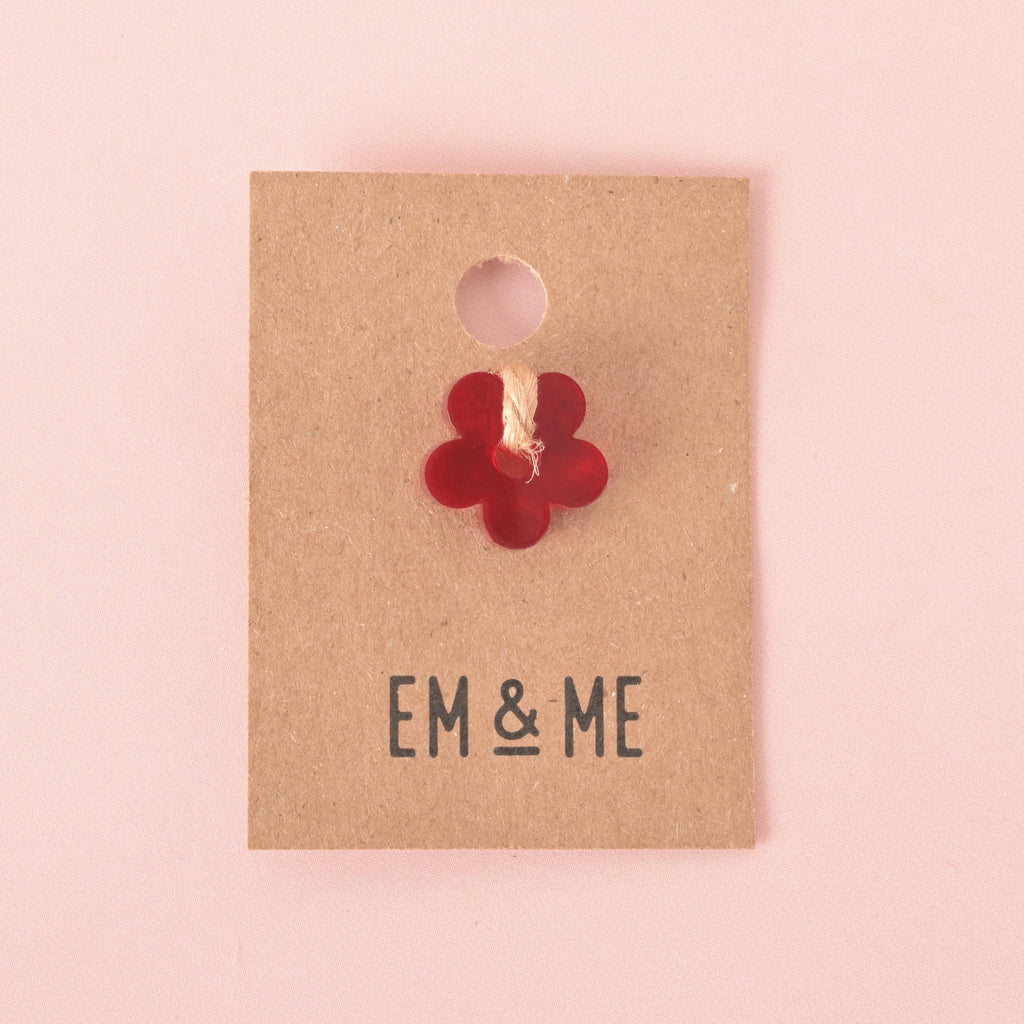 A single red, flower-shaped mini charm attached to a brown paper card that reads "EM & ME." The item is displayed on a solid pink background.