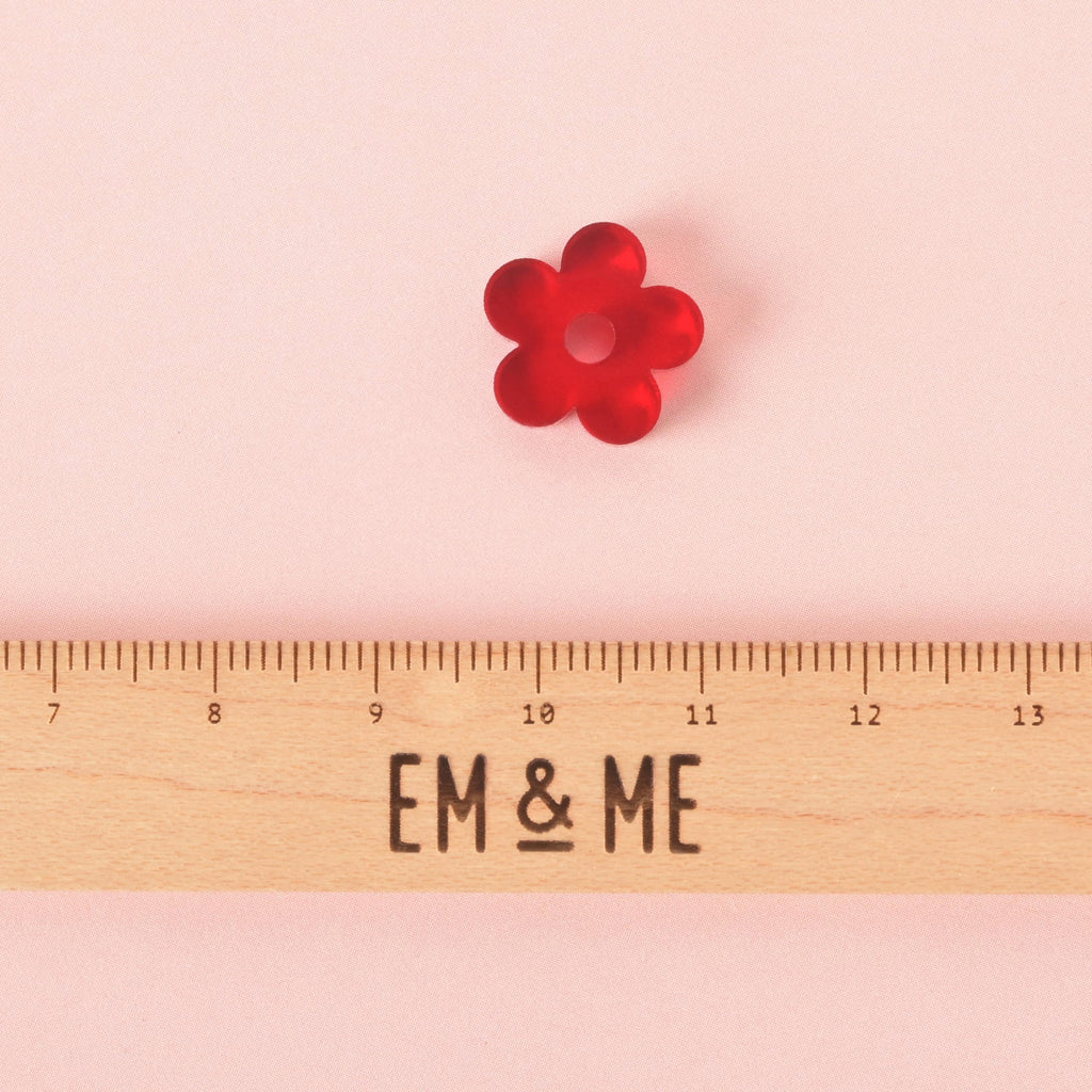 A single red, flower-shaped charm with a hole in the middle. A wooden ruler with the "EM & ME" logo is placed below the charm to show its size. The charm is shown on a solid pink background.