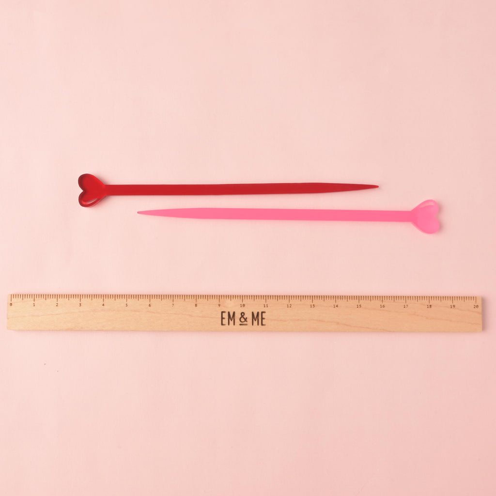 Two acrylic stir sticks with heart-shaped tips, one in red and one in light pink, lying horizontally. They are displayed above a wooden ruler marked with the brand EM & ME, on a solid light pink background.