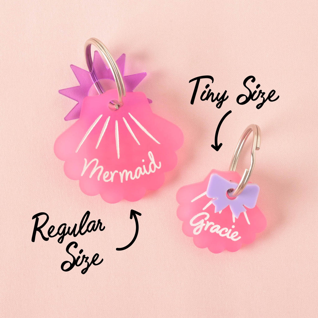 Two pink seashell-shaped pet tags are shown for size comparison. The larger tag says "Mermaid" and has purple star charms, while the smaller tag says "Gracie" and has a purple bow charm. The image includes the text "Regular Size" and "Tiny Size" with arrows pointing to each tag. The items are displayed on a solid pink background.