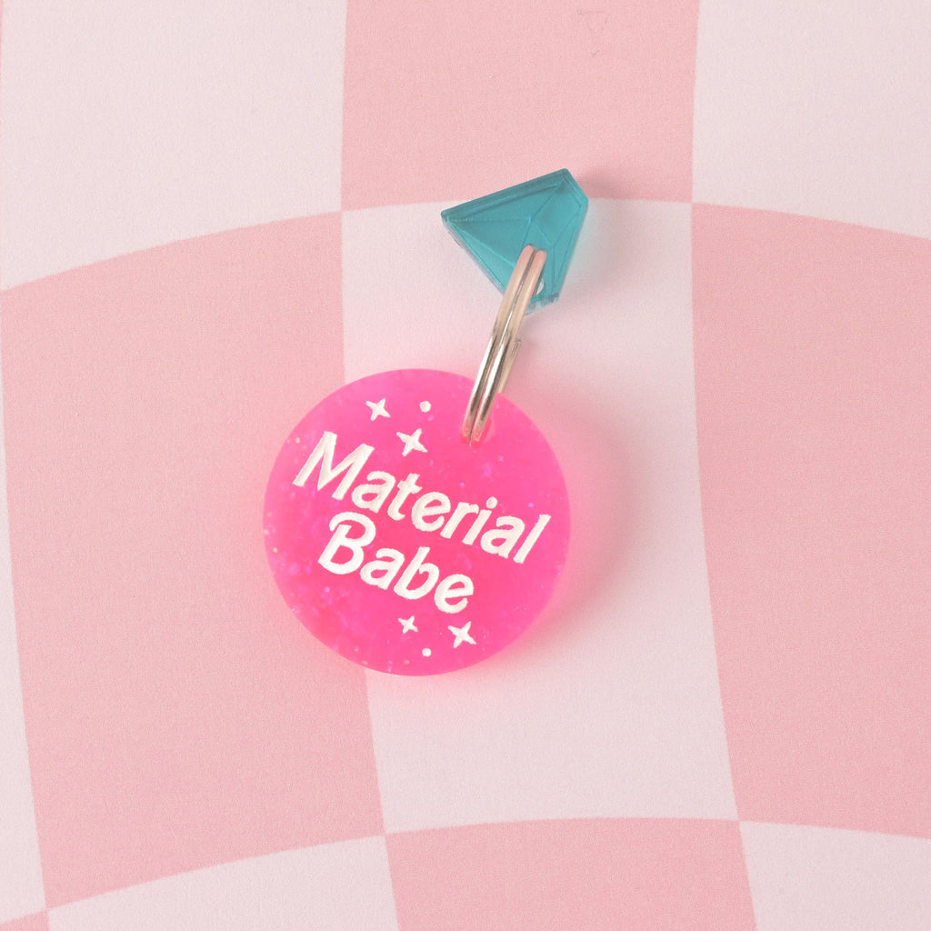 A round acrylic tag made from vibrant hot pink glitter, engraved with the white slogan "Material Babe" and star graphics. The tag is accented with a turquoise diamond-shaped gemstone charm and attached to a split ring. Material Babe, Barbie Inspired Personalized Pet Tag, ID Tag for Cats and Dogs, Limited Edition, Custom Name and Contact Info Engraved