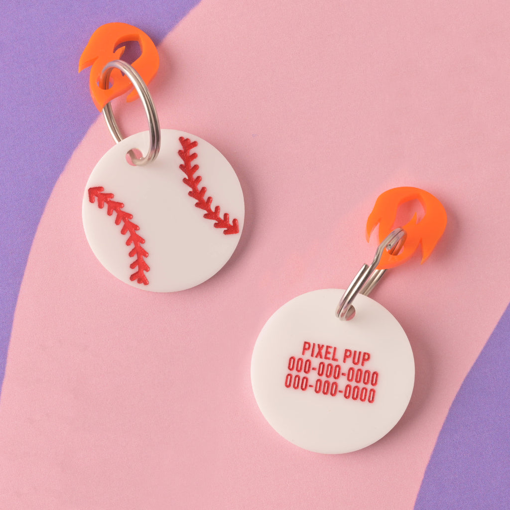 Two round white acrylic pet ID tags shaped like a baseball with red stitching. One shows the baseball design, and the other shows personalization details (PIXEL PUP and phone numbers). Both tags have an orange acrylic clip/charm and are displayed on a purple and pink striped background.