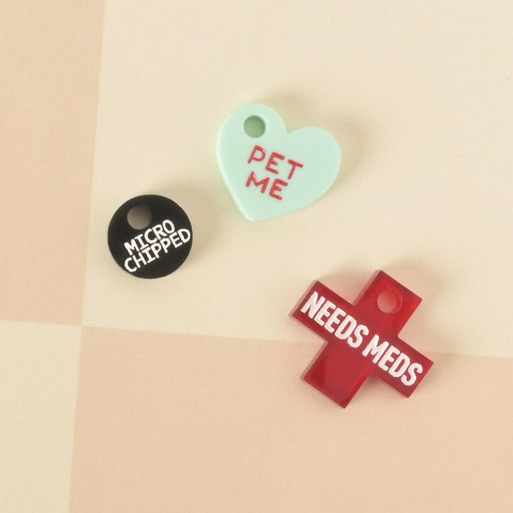 A set of three pet charms. One is a black circle that says "MICROCHIPPED," one is a mint green heart that says "PET ME," and one is a red cross that says "NEEDS MEDS." The items are displayed on a light yellow and white checkered background.