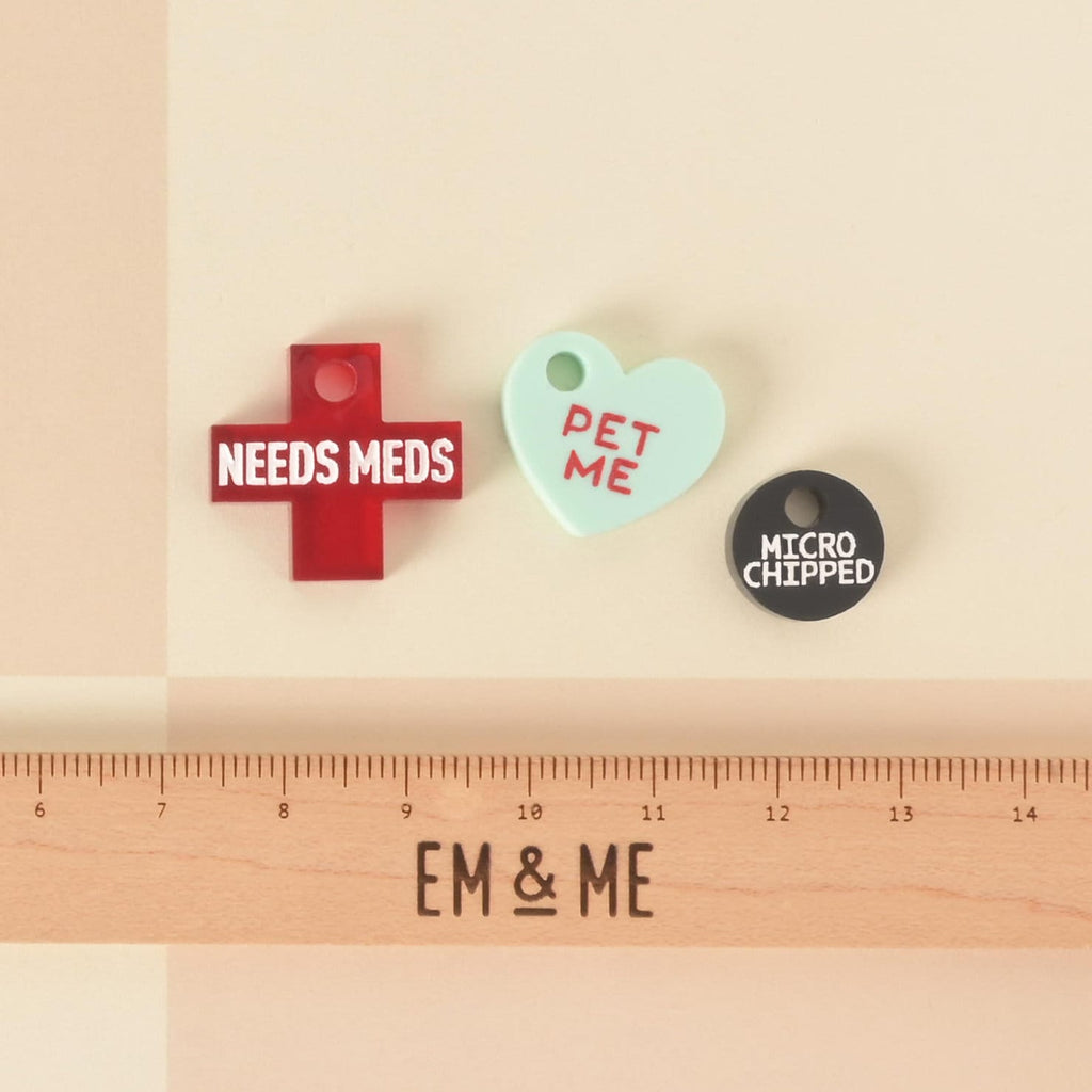 A set of three pet charms. One is a red cross that says "NEEDS MEDS," one is a mint green heart that says "PET ME," and one is a black circle that says "MICROCHIPPED." A wooden ruler with the "EM & ME" logo is placed below the charms to show their size. The items are shown on a light yellow and white checkered background.
