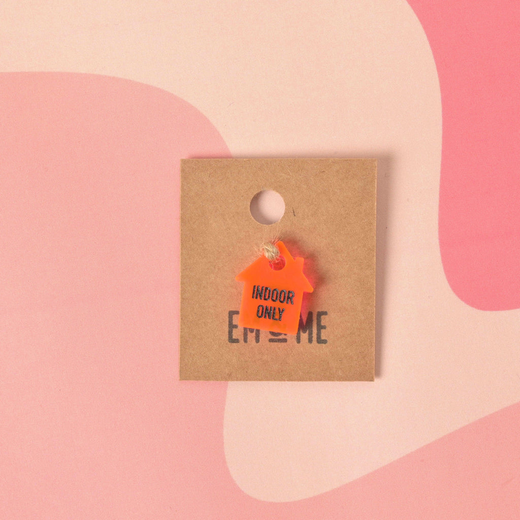 Close-up of a small, fluorescent pink acrylic charm shaped like a house, engraved with the text "INDOOR ONLY." The charm is attached to a string and displayed on a square kraft paper backing card branded with "EM & ME." Indoor Only, House Cat Seasonal Mini - emandmestudio