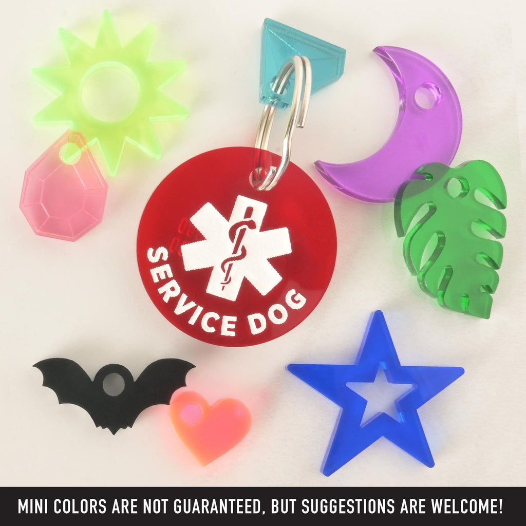 A product photo of a round, red pet ID tag with a white medical symbol and the words "SERVICE DOG" around the bottom edge. Surrounding the main tag are several smaller, colorful accessories: a neon green sun, a pink faceted gem, a black bat, a light orange heart, a blue star, a green monstera leaf, and a purple crescent moon. Text at the bottom of the image reads, "MINI COLORS ARE NOT GUARANTEED, BUT SUGGESTIONS ARE WELCOME!"