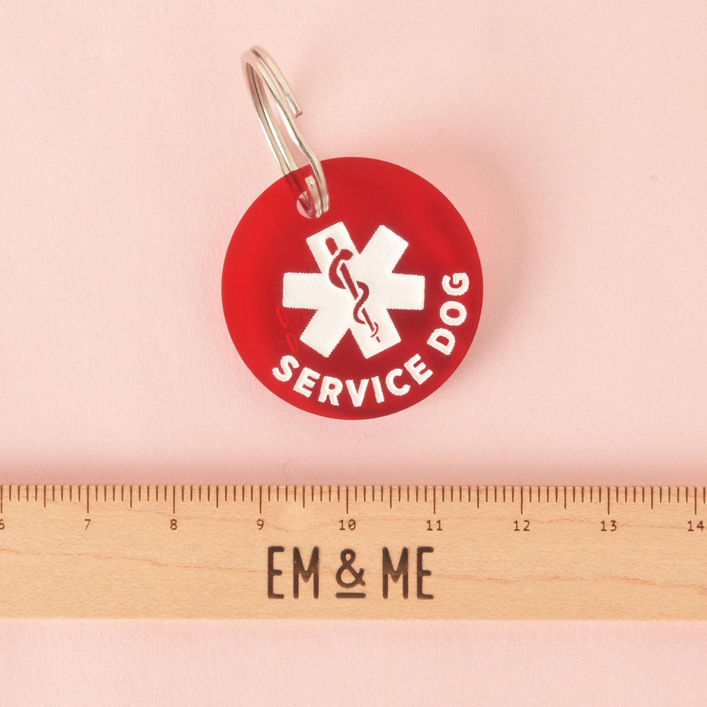 A product photo of a round, red pet ID tag with a white medical cross symbol and the words "SERVICE DOG" around the bottom edge. The tag is shown on a pink background next to a silver key ring. A wooden ruler with the words "EM & ME" printed on it is positioned below the tag to show its size.