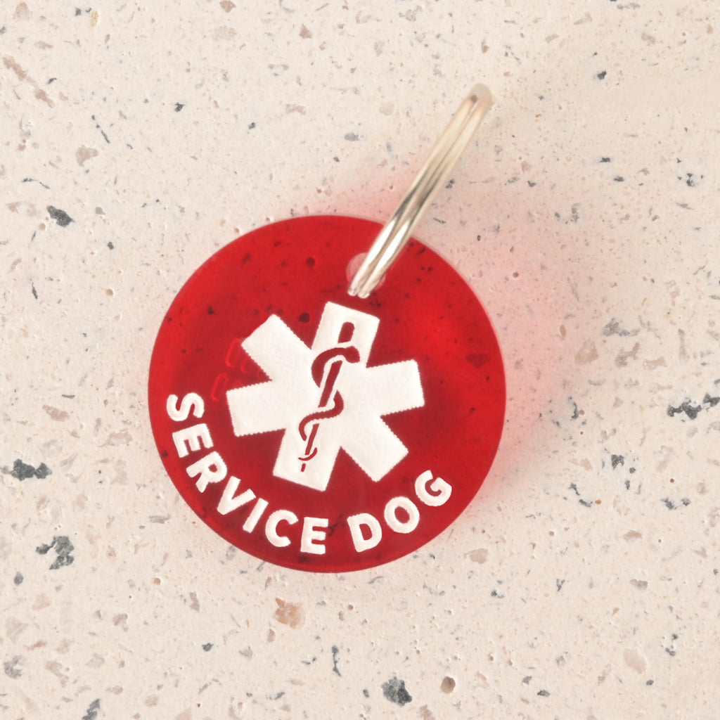 A close-up product photo of a round, translucent red pet ID tag with a white medical symbol and the words "SERVICE DOG" engraved on it. The tag is shown on a light, speckled surface and includes a silver split key ring.