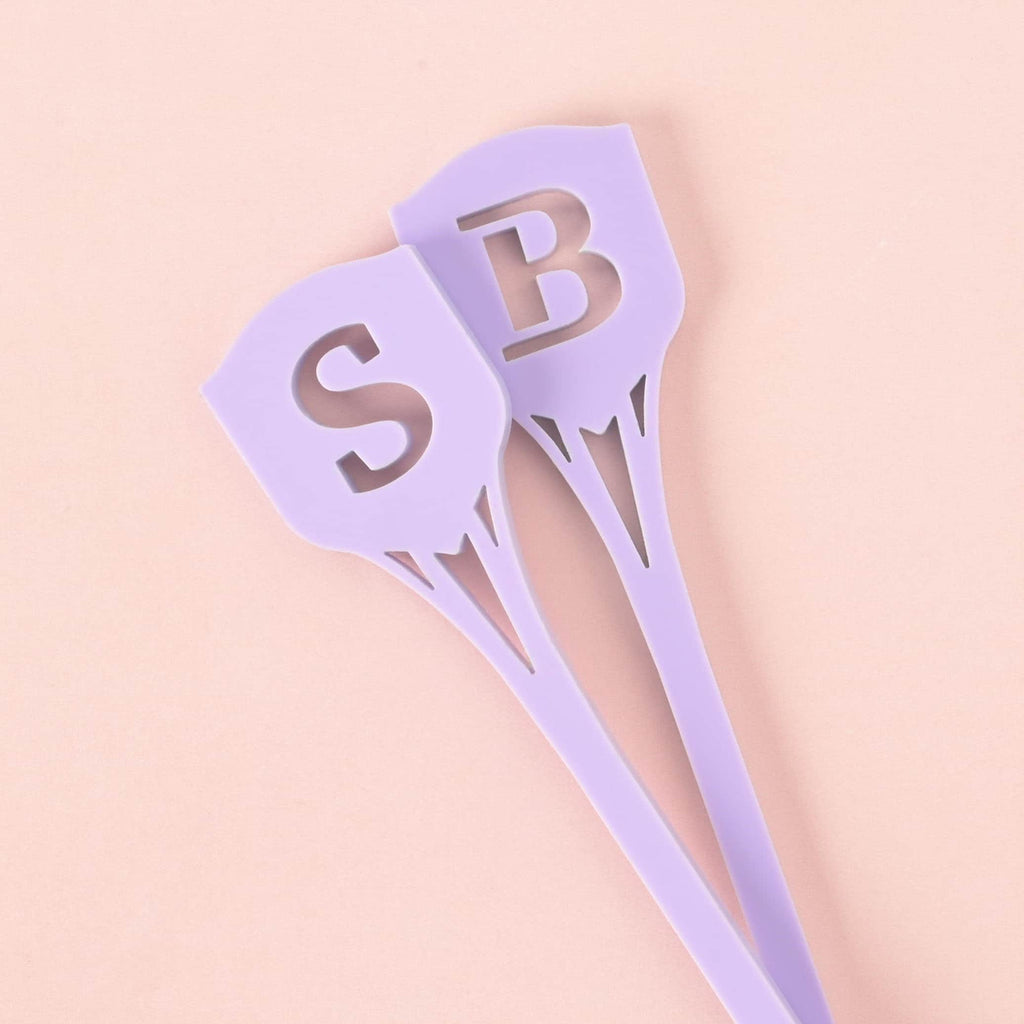 A close-up of two solid purple cocktail stirrers. The stirrers feature the cutout monogram letters 'S' and 'B' at the top in a shield shape, overlapping each other on a solid pink background.