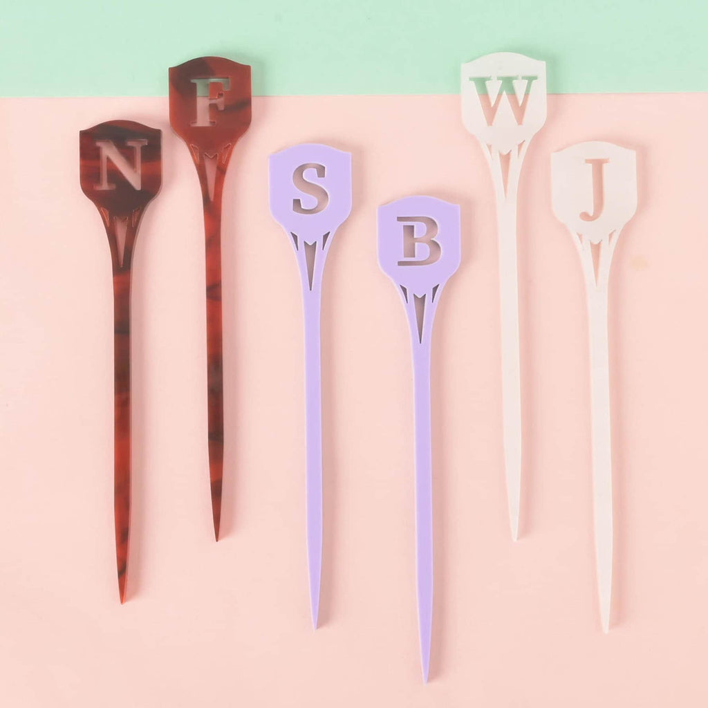 A set of six personalized monogram cocktail stirrers, arranged on a pink and mint green background. The stirrers include two in red tortoiseshell with the letters 'N' and 'F', two in solid purple with 'S' and 'B', and two in white iridescent with 'W' and 'J'. All stirrers have a shield shape at the top with a pointed bottom.