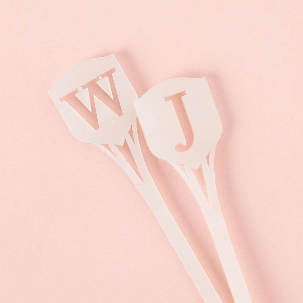 A close-up of two white iridescent cocktail stirrers. The stirrers feature the cutout monogram letters 'W' and 'J' at the top in a shield shape, overlapping slightly on a solid pink background.