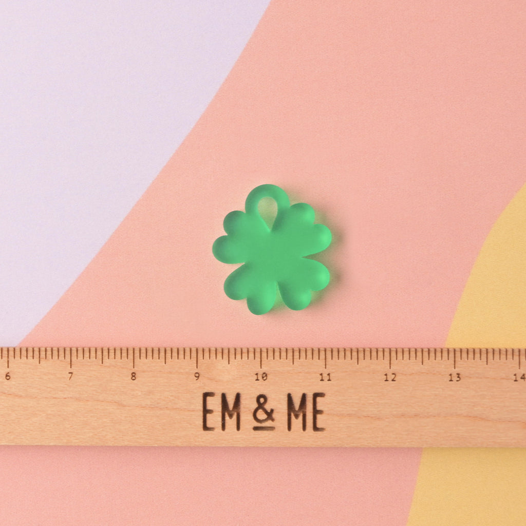 Close-up of a single, matte medium-green acrylic charm shaped like a four-leaf clover. The charm is displayed above a wooden ruler marked with the brand EM & ME, on a color-blocked background of pink, purple, and white.