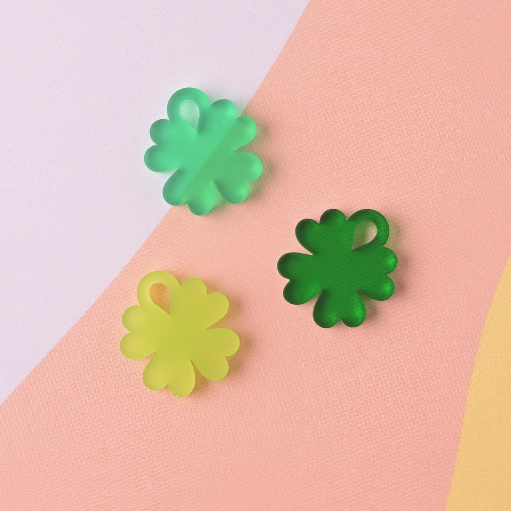 Three small acrylic charms shaped like four-leaf clovers, displayed on a pink, purple, and white striped background. The charms are in three different shades of green: neon/lime green, translucent medium green, and translucent dark green.