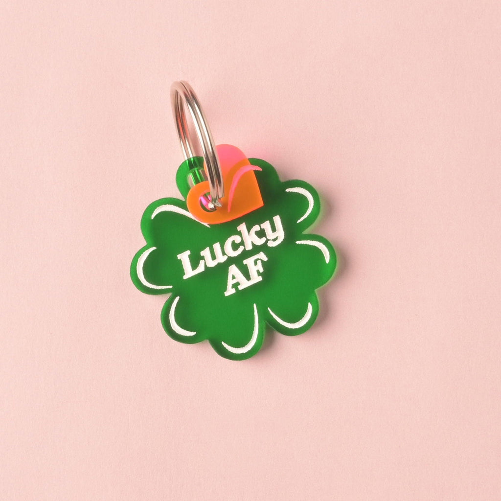 Close-up of a translucent green, clover-shaped acrylic pet ID tag featuring the white, humorous slogan 'Lucky AF.' A small pink and orange layered heart charm is attached to the silver split ring, displayed on a pink background. Lucky AF, 4 leaf Clover Personalized Pet Tag, St Patrick Cat and Dog ID Tag