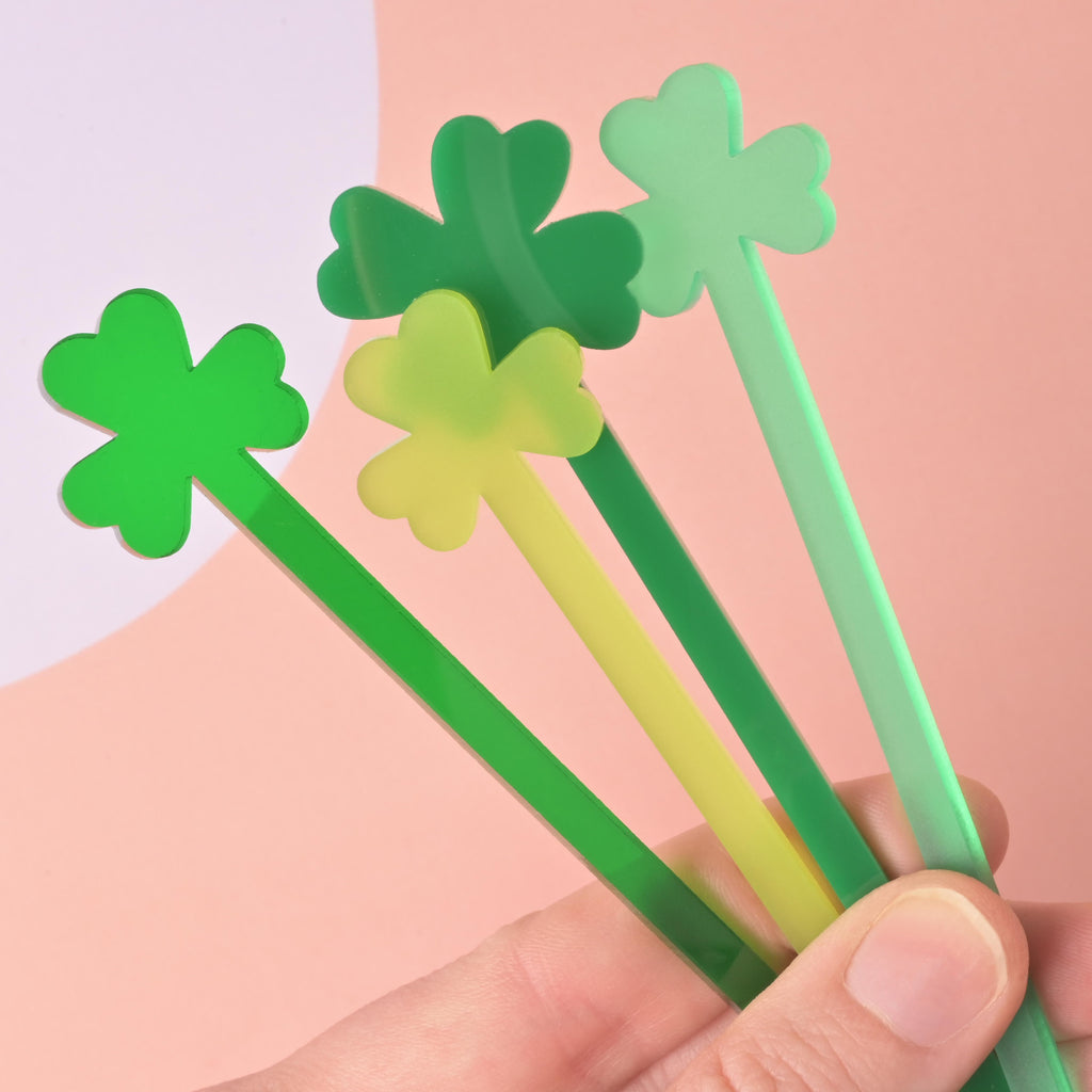 A hand holding a fanned group of four acrylic stir sticks with clover-shaped tops. The sticks are in various shades of green, including lime, medium green, and dark green. They are held against a pink and white background.