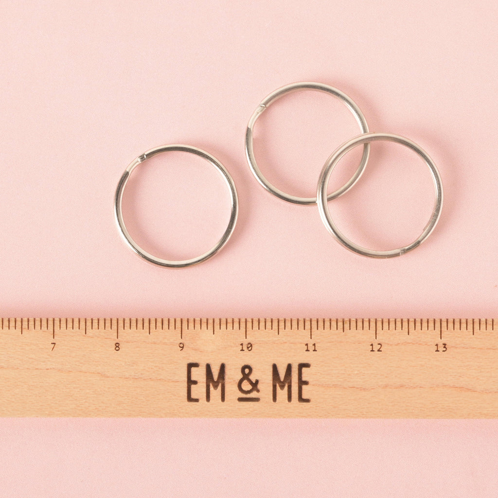 A product photo showing three silver split key rings, positioned above a wooden ruler with the words "EM & ME" to show scale. The key rings and ruler are on a pink background.