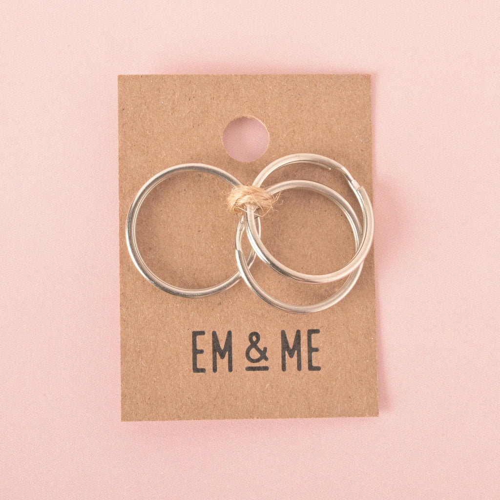 A product photo showing three silver split key rings of different sizes, tied together with twine on a brown cardboard display card. The card has a circular cutout at the top and the words "EM & ME" printed at the bottom. The items are on a pink background.