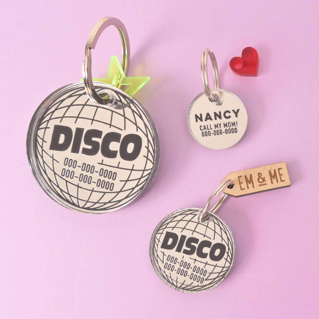 A collection of three personalized pet tags and a small red heart charm. The tags include a large disco ball that says "DISCO" with a placeholder phone number, a smaller disco ball tag that reads "EM & ME" on an attached wooden charm, and a white round tag that says "NANCY CALL MY MOM!" The tags are attached to silver rings and are displayed on a solid pink background.