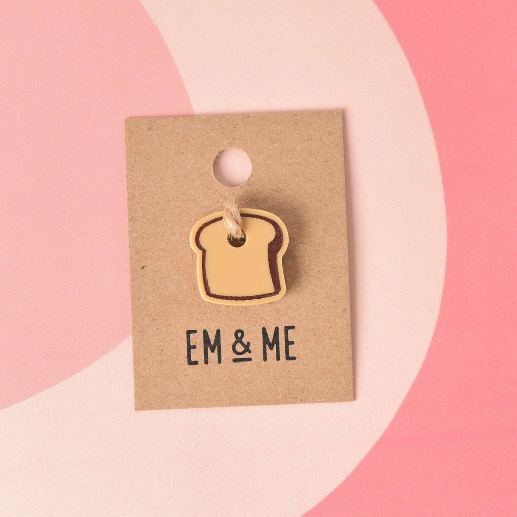 A small, layered acrylic charm shaped like a slice of toast with a light brown/tan center and a darker crust outline. The charm is attached to a string and displayed on a small square kraft paper backing card branded with "EM & ME." Bread Mini Charm, Tiny Toast Accessory for Pet ID Tag, Cats and Dogs Add-on Charm