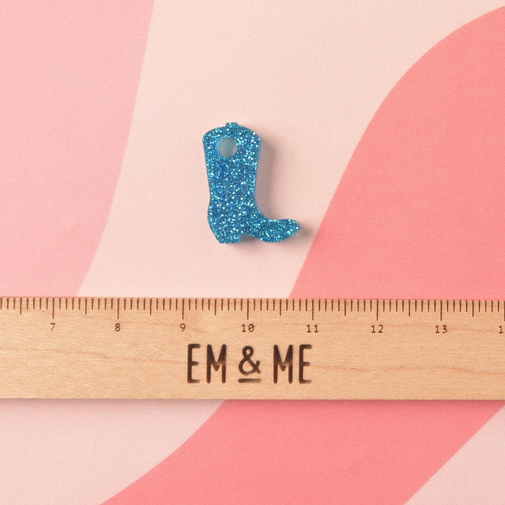 A small, laser-cut acrylic mini charm shaped like a cowboy boot in vibrant blue glitter. The charm is shown above a wooden ruler with the 'EM & ME' logo to indicate its size.