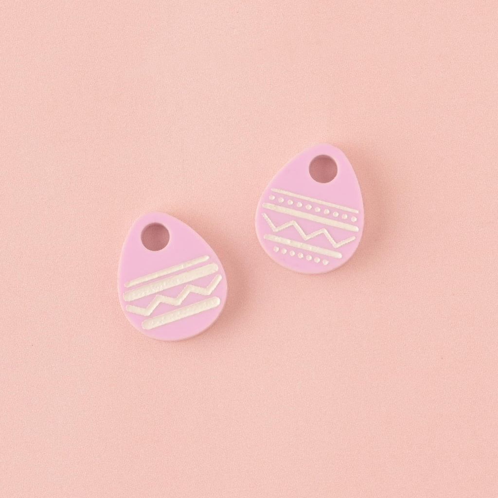 Two small, light purple (lilac) acrylic charms shaped like Easter eggs, both decorated with white engraved zigzag and stripe patterns. The charms are displayed side-by-side on a solid light pink background.