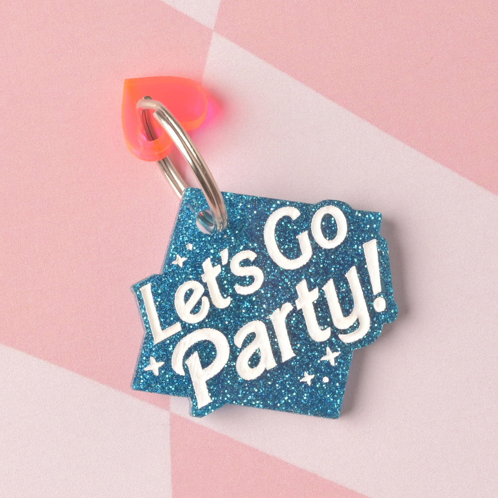 A custom acrylic tag shaped like a diamond or banner in vibrant blue glitter, engraved with the white slogan "Let's Go Party!" The tag is accented with a small iridescent pink and orange heart charm on a split ring. Barbie Inspired Personalized Pet Tag, Let's Go Party ID Tag for Cats and Dogs, Limited Edition, Custom Name and Contact Info Engraved