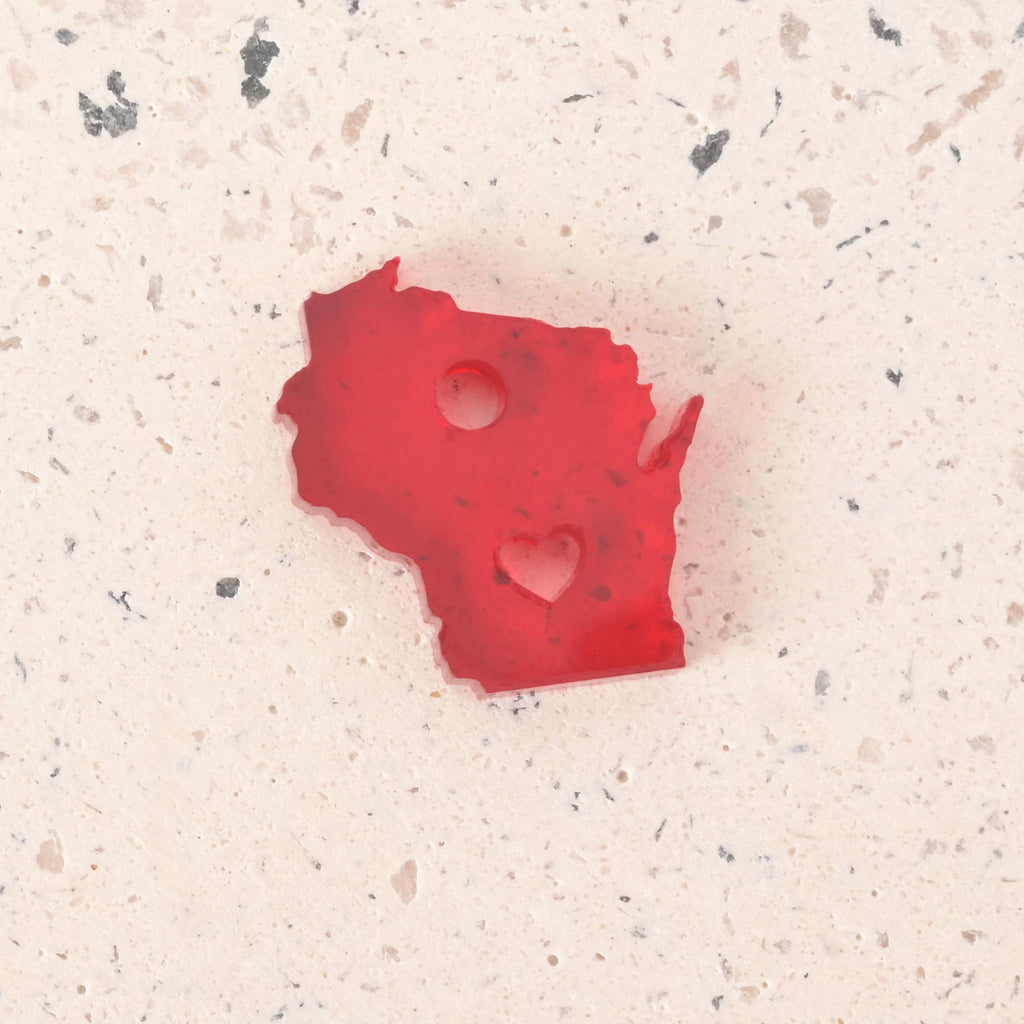 Speckled red acrylic Wisconsin state-shaped charm with heart cutout and top hanging hole, placed on a light terrazzo background.