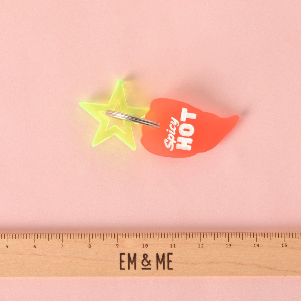 Close-up of a translucent orange/red chili pepper-shaped pet ID tag with the white slogan "Spicy HOT." The tag is attached to a layered neon yellow-green star charm and displayed above a wooden ruler marked with the brand EM & ME. Spicy Hot, Pepper Personalized Pet Tag, ID for Cats and Dogs, Veggie Pet Tags