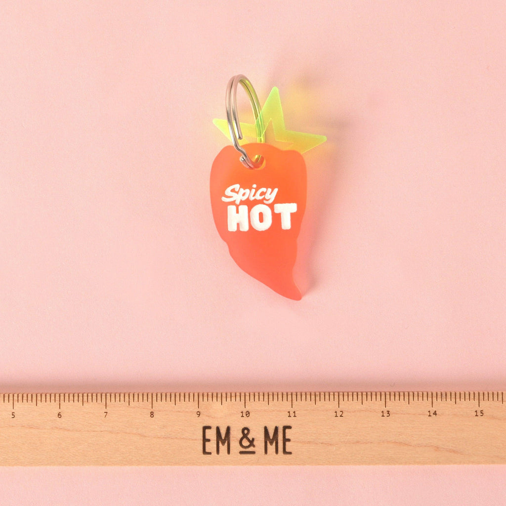 Close-up of a translucent orange/red chili pepper-shaped pet ID tag with the white slogan "Spicy HOT." The tag is attached to a neon yellow-green charm and displayed above a wooden ruler marked with the brand EM & ME. Spicy Hot, Pepper Personalized Pet Tag, ID for Cats and Dogs, Veggie Pet Tags
