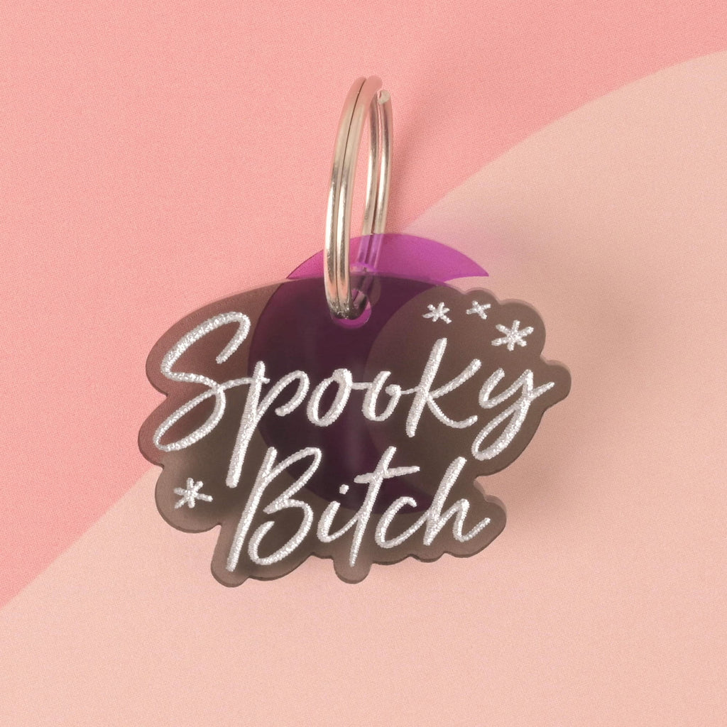 A close-up product photo of a gray, translucent pet tag with a unique cloud or ghost-like shape. The tag is engraved with the words "Spooky Bitch" in white and has small star accents. A purple geometric charm is attached to the top of the tag, which is shown with a silver split key ring.