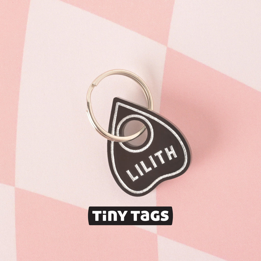 Close-up of a single black planchette-shaped pet ID tag with the name LILITH engraved in white, attached to a silver split ring, set against a pink and white checkered background. The brand TINY TAGS is visible below the tag. Small Ouijia Planchette Pet tag, Tiny Tag, Spooky Halloween Cat or small dog breeds, XS personalized pet name tag
