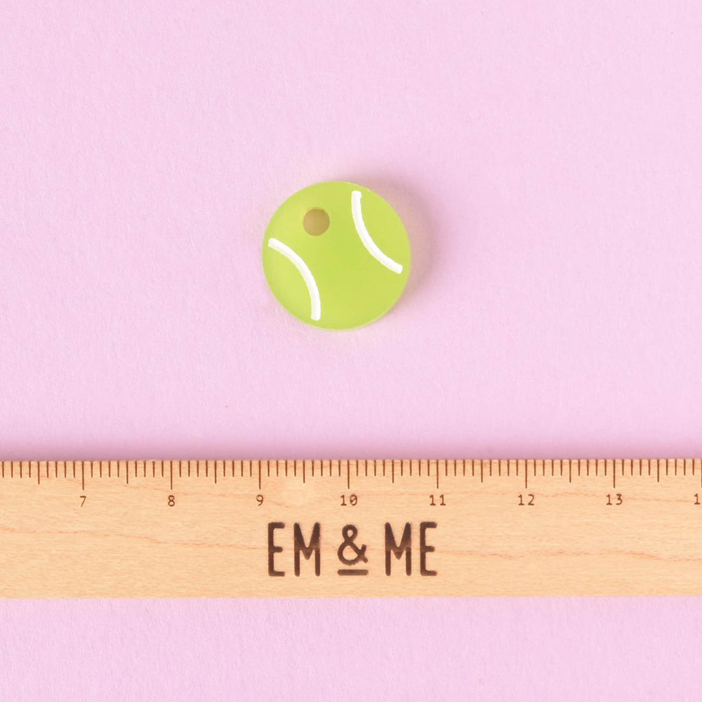 A close-up of a small, bright green tennis ball-shaped charm or mini pet tag with white curved lines. A wooden ruler with the "EM & ME" logo is placed below the charm to show its size. The item is displayed on a solid pink background.

