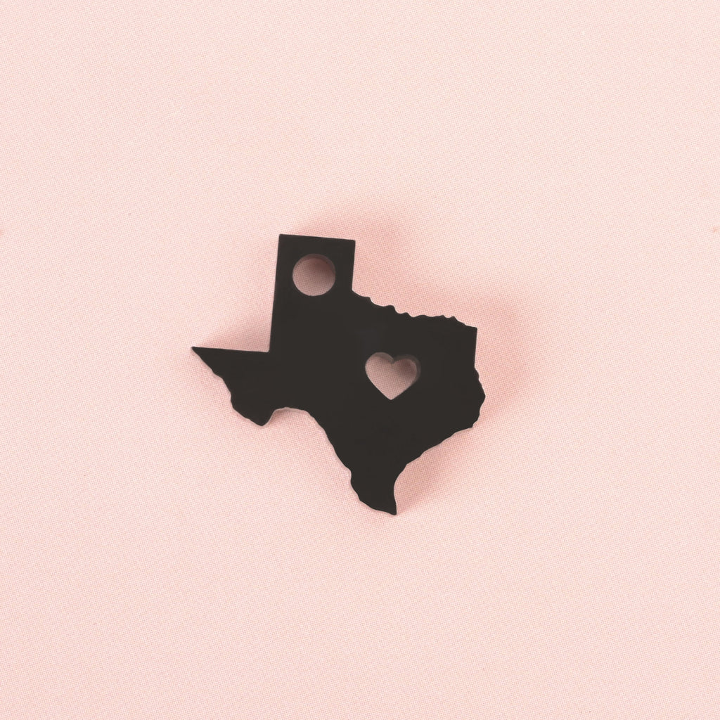 Black acrylic Texas state-shaped charm with a heart cutout and top hole for hanging, displayed on a soft pink background.
