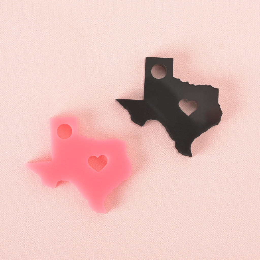 Two acrylic Texas state-shaped charms with heart cutouts, one in black and one in pink, displayed on a soft pink background.