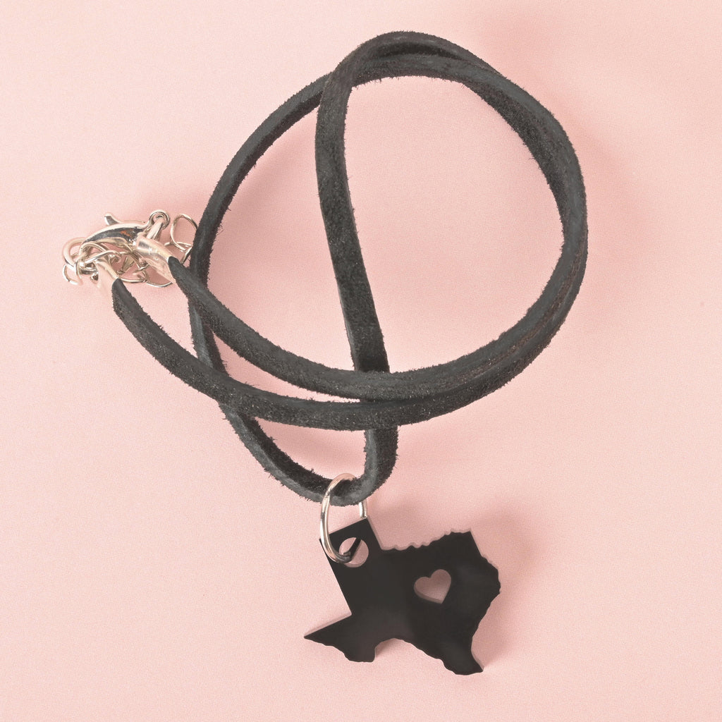 Texas state-shaped black acrylic pendant with heart cutout, hanging from a black faux suede cord necklace with silver clasp, displayed on a pink background.